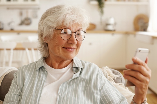 happy joyful female pensioner round eyeglasses surfing internet cell phone looking mobile s screen with broad smile booking plane tickets planning trip scrolling pics via social network